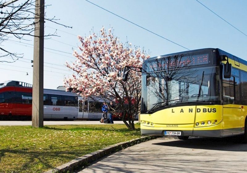 Ein Landbus steht an einer Haltestelle bei einem Bahnhof, im Hintergrund sind ein blühender Baum und ein Zug zu sehen.