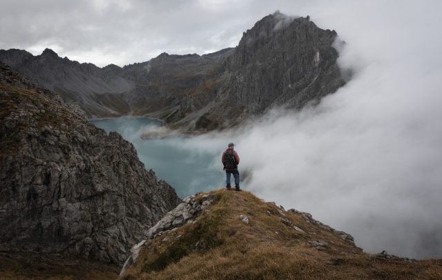 Eine Person steht auf einem Berggipfel und schaut in die umliegende Bergwelt, im Tal ist der Lünersee zu sehen, Nebelz zieht auf.