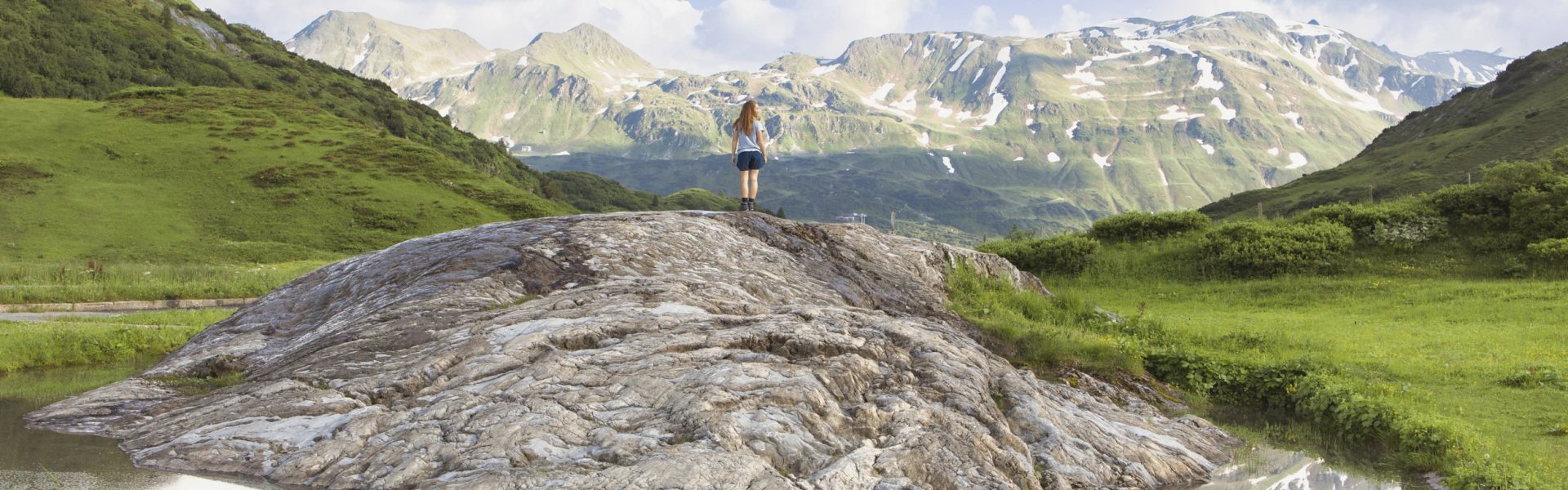 Gebirgslandschaft in Lech am Arlberg mit kleinem Gewässer und Fels im Vordergrund, Wanderin und Hochgebirgslandschaft im Hintergrund.