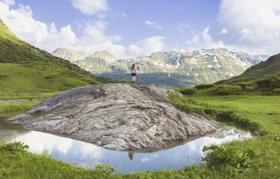 Gebirgslandschaft in Lech am Arlberg mit kleinem Gewässer und Fels im Vordergrund, Wanderin und Hochgebirgslandschaft im Hintergrund.