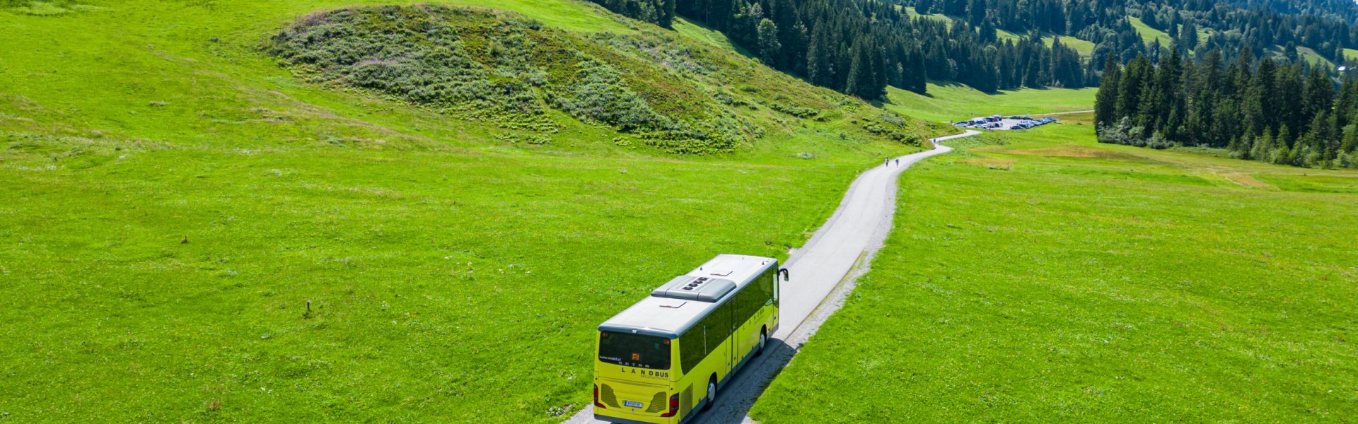 Ein Landbus fährt durch eine sommerliche Landschaft mit grünen Wiesen, Wäldern und Bergen nach Schönenbach im Bregenzerwald.