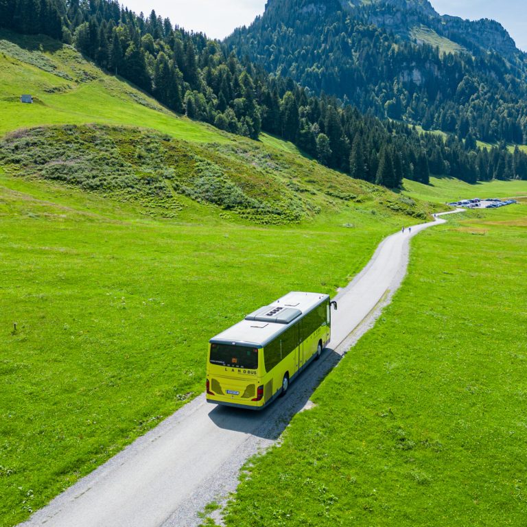 Ein Landbus fährt durch eine sommerliche Landschaft mit grünen Wiesen, Wäldern und Bergen nach Schönenbach im Bregenzerwald.