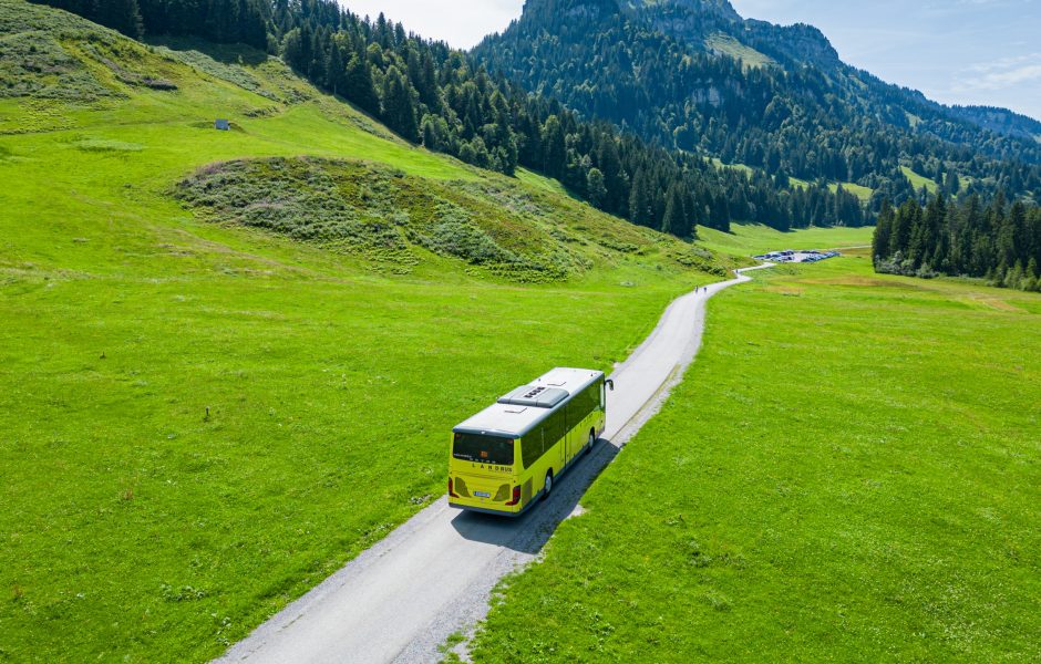 Ein Landbus fährt durch eine sommerliche Landschaft mit grünen Wiesen, Wäldern und Bergen nach Schönenbach im Bregenzerwald.