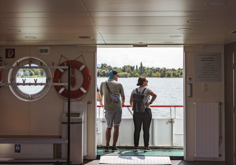 Blick vom Innenraum eines Schiffes der Vorarlberg Lines auf eine Frau und einen Mann, die auf den Bodensee und das Bodenseeufer schauen.