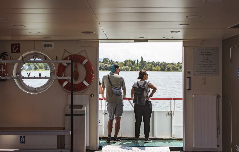Blick vom Innenraum eines Schiffes der Vorarlberg Lines auf eine Frau und einen Mann, die auf den Bodensee und das Bodenseeufer schauen.