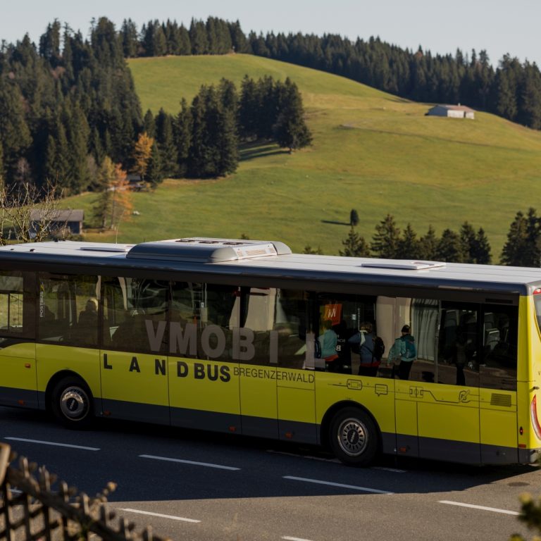 Ein Landbus in Vorarlberg ist im Vordergrund zu sehen, im Hintergrund hügelige Landschaft mit Wiesen und Wäldern.
