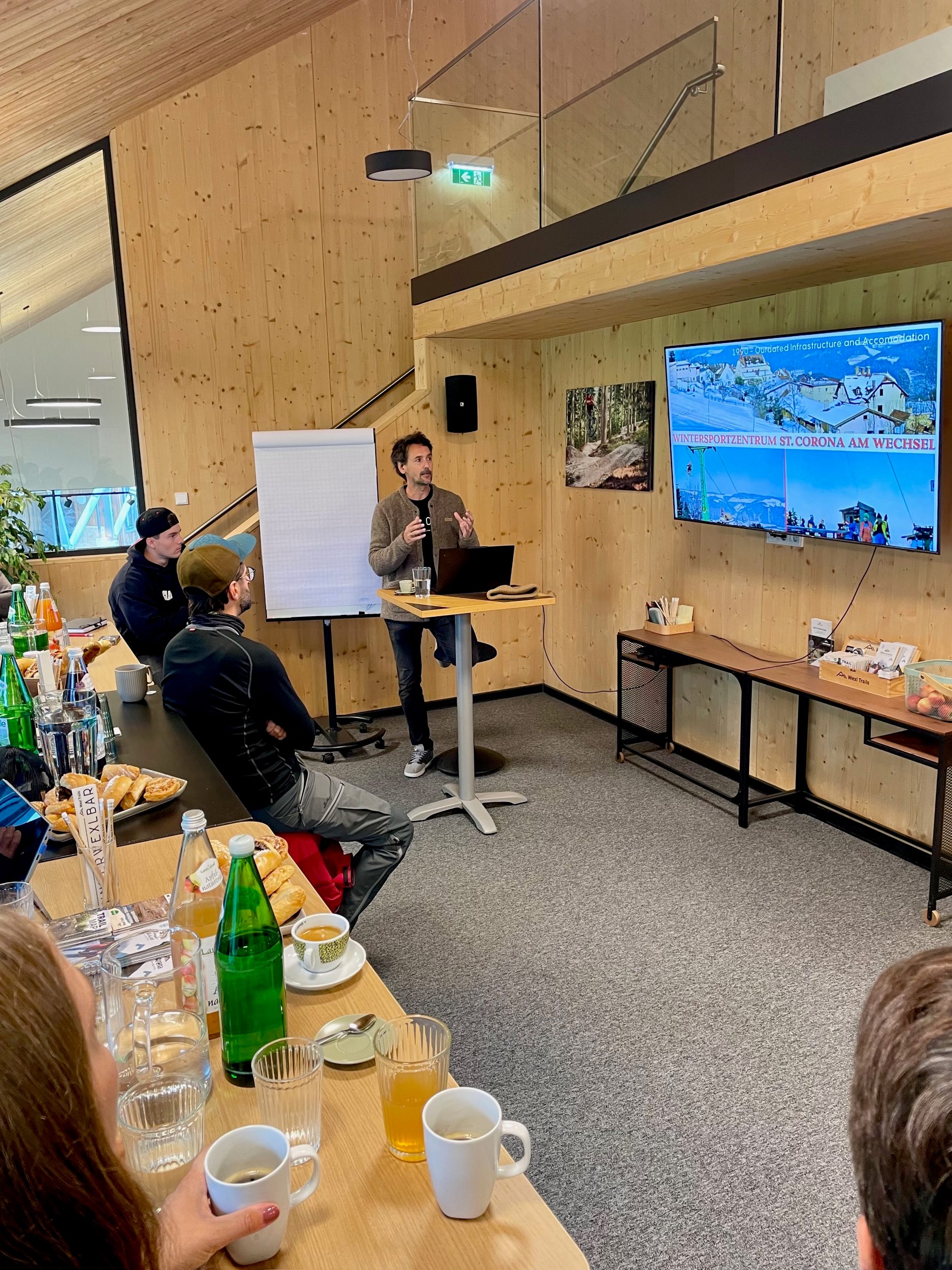 Eine vortragende Person mit einer Präsentation in einem offenen, holzigen Raum, fotografiert aus der Zuhörer:innenperspektive.