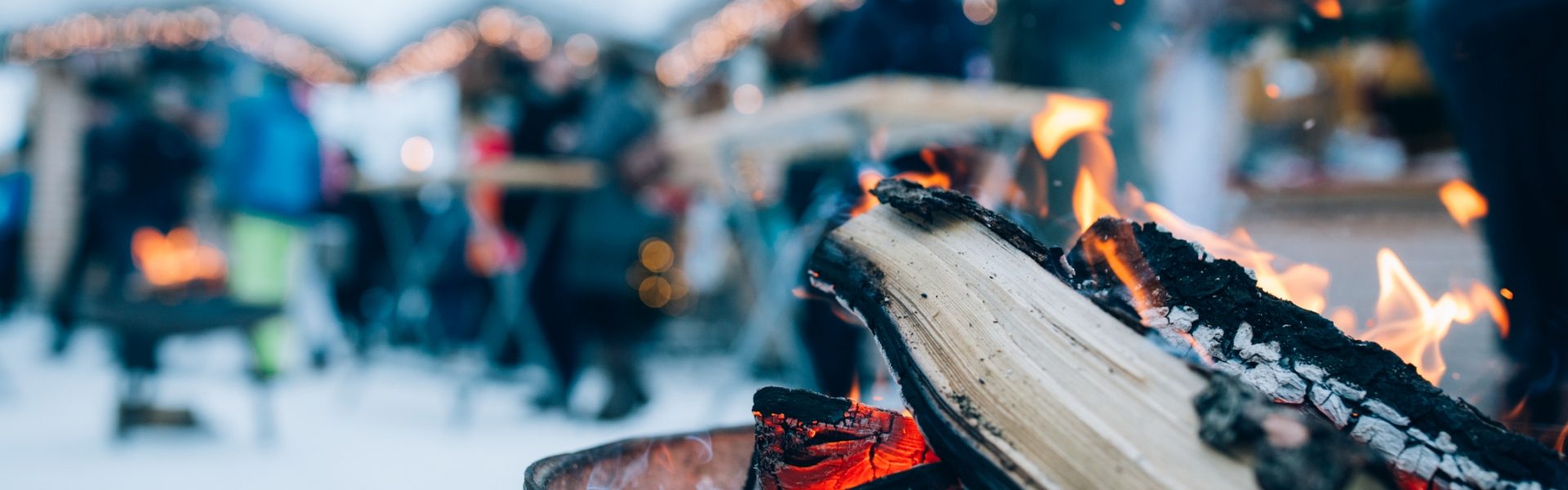 Adventstimmung mit Feuerschale im Vordergrund und Christkindlmarkt in verschneiter Landschaft im Hintergrund.