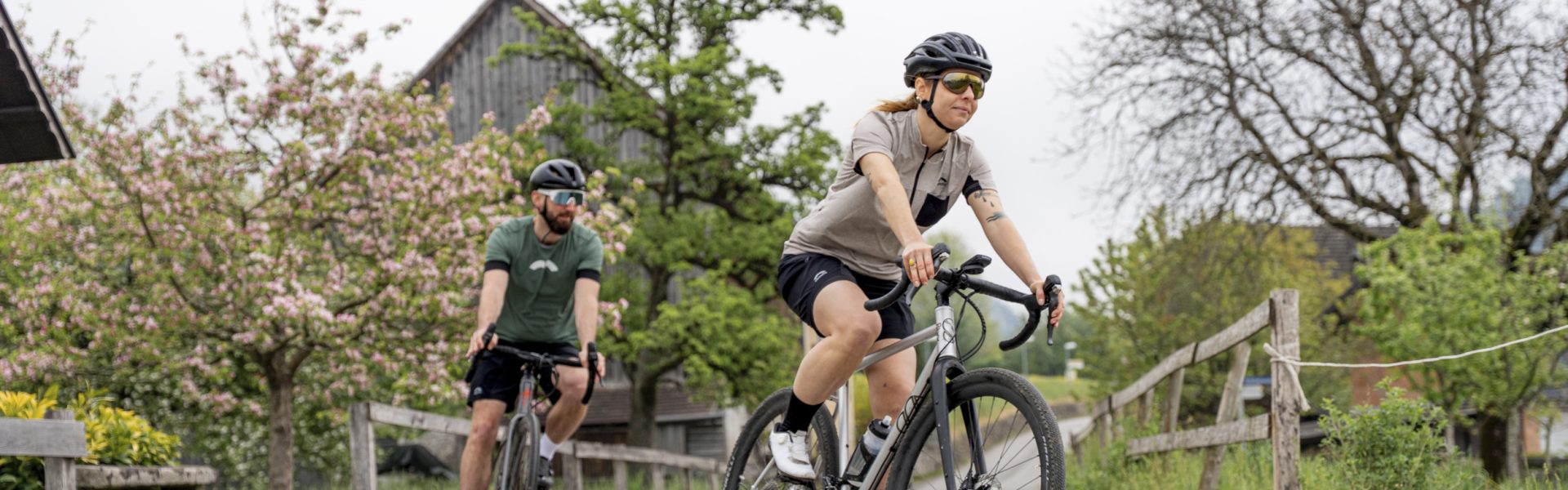 Zwei Radfahrer:innen fahren auf einer ruhigen Straße durch die blühende Kulturlandschaft bei Bludenz in Vorarlberg, vorbei an Obstbäumen, Holzzäunen und ländlicher Bebauung. - Gravelbike Bludenz (c) Chris Gollhofer - Vorarlberg Tourismus