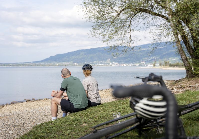Zwei Radfahrer:innen sitzen am Ufer des Bodensees in Vorarlberg.