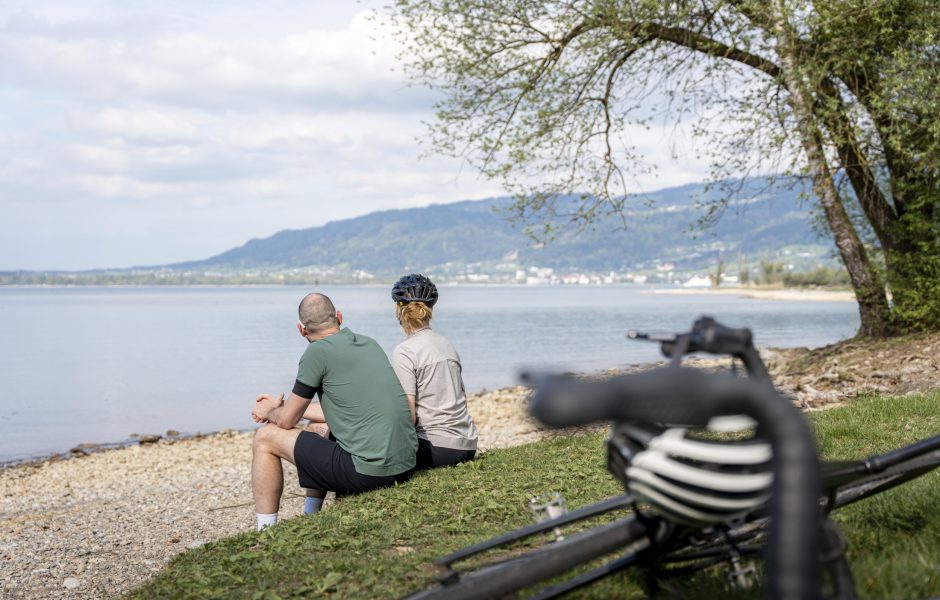 Zwei Radfahrer:innen sitzen am Ufer des Bodensees in Vorarlberg.