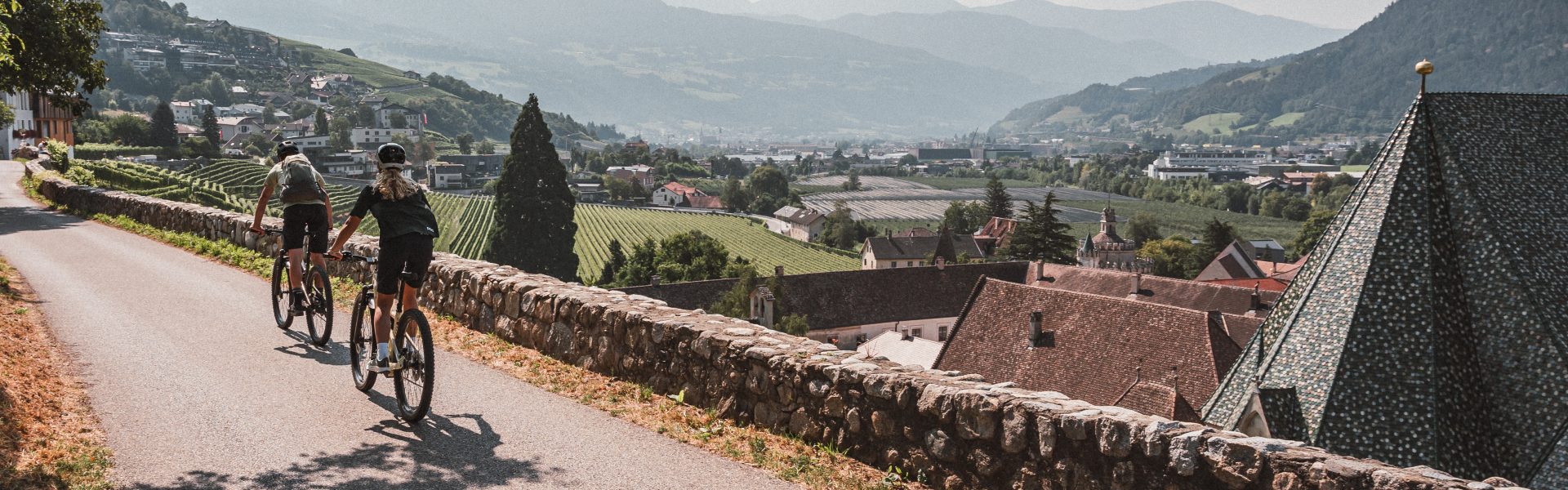 Zwei Radfahrende auf einer Straße oberhalb von Brixen mit Blick auf Weinberge, Altstadt und die umliegenden Berge in Südtirol.